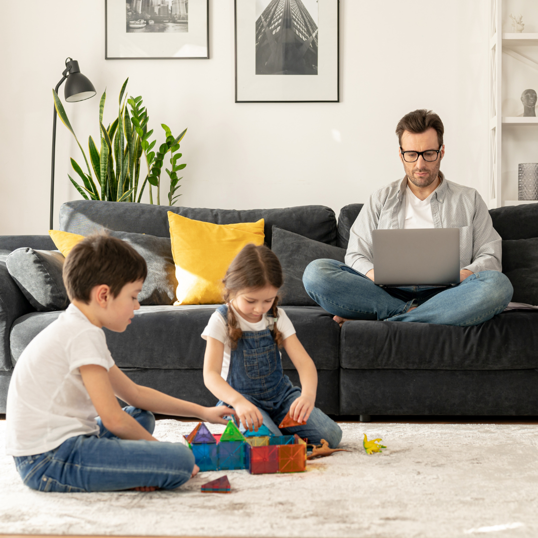 A man with computer working while his kids are playing on the carpet with blocks. The man could be looking for financial planning tips for newly single parents.